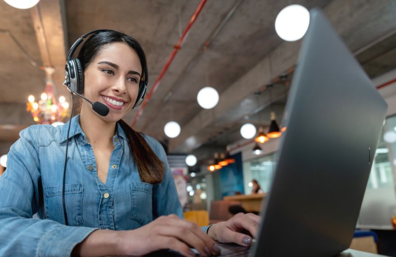 Mujer con auriculares trabajando en asistencia virtual desde una laptop en una oficina moderna.